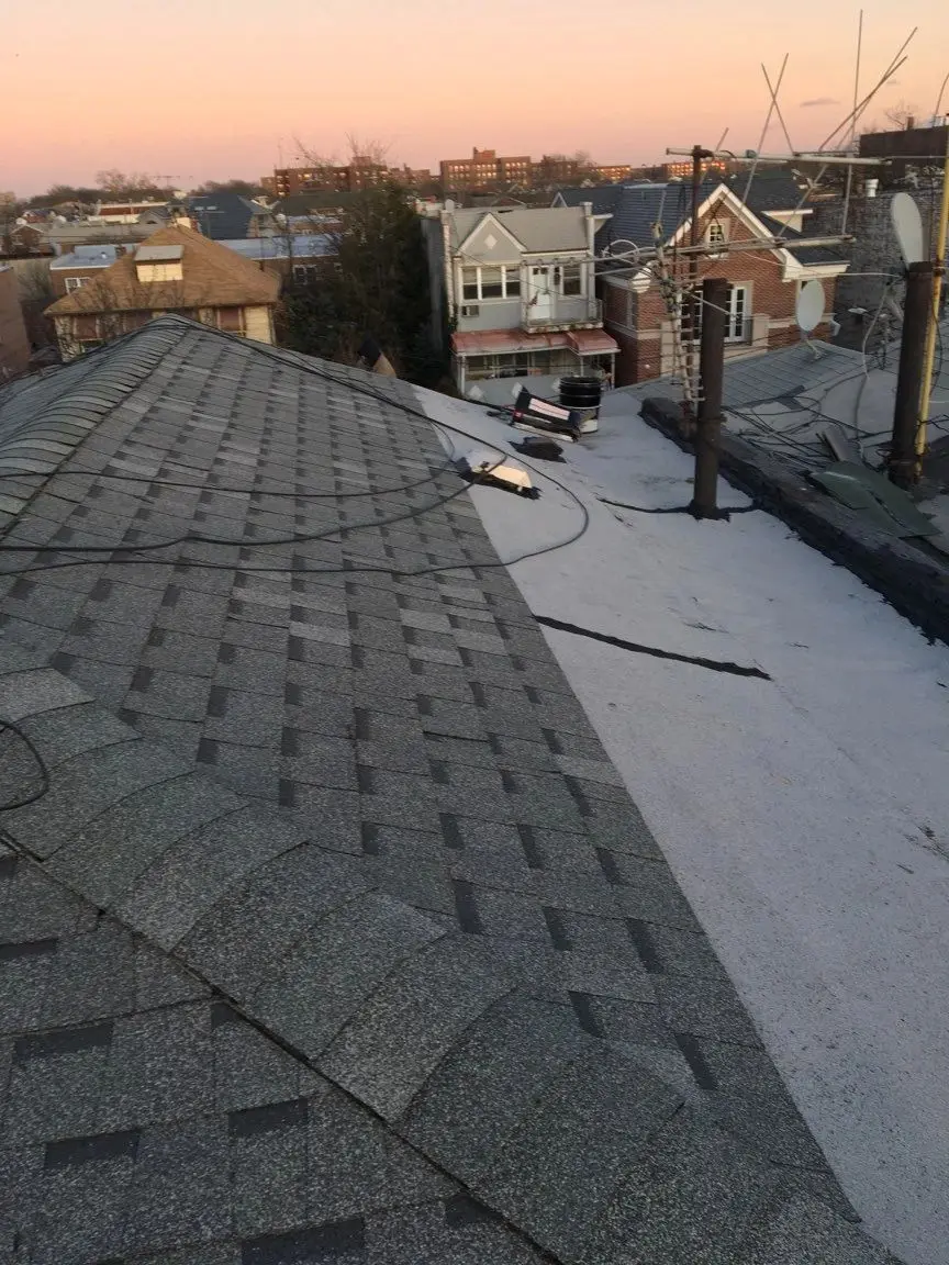 Shingle roof meeting a flat roof section at dusk, with neighboring houses behind